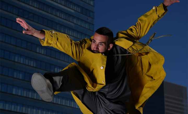 Con edificios urbanos de fondo, un hombre de cabello corto con una chaqueta amarilla es fotografiado en el momento de un salto.