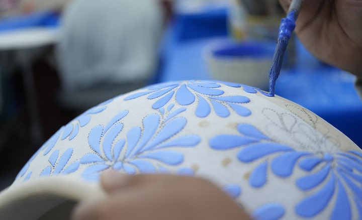 Imagen que muestra a un artesano añadiendo patrones a una jarra con un pincel y pintura azul. El enfoque está en el pincel, y el primer plano de la jarra y el fondo están desenfocados